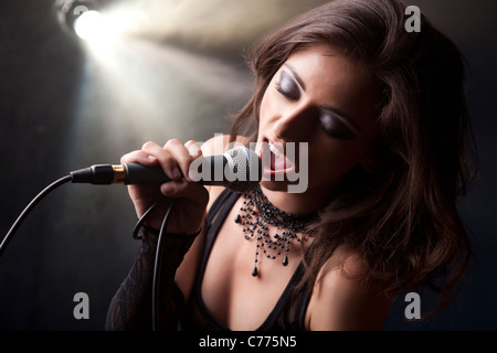 Giovane e bella ragazza a cantare in un atmosfera di roccia che indossa un top nero, nero Guanti di pizzo e di girocollo perle su un livello di grigio smokey indietro Foto Stock