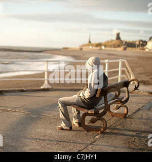 Un giovane uomo che indossa una felpa con cappuccio da sola seduta su una panchina sul mare, Aberystwyth Wales UK Foto Stock