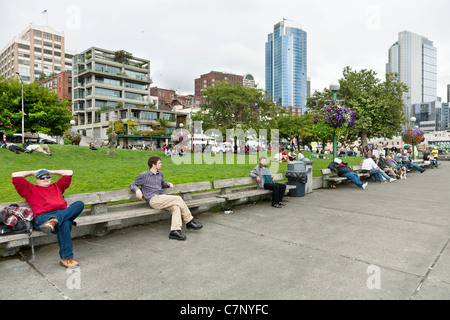 Le persone amano sedere & giacente sulla fila di panche o sollevato prato di Victor Steinbrueck Park contro il fondale di Urban Skyline Foto Stock