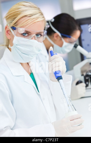 Una bionda femmina o medico ricercatore scientifico o medico usando la sua pipetta in un laboratorio con il suo collega e un microscopio Foto Stock