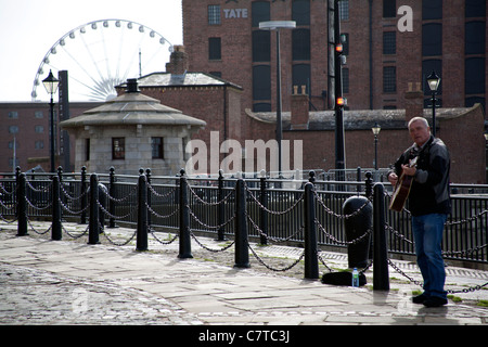 Un suonatore ambulante di suonare una chitarra su Albert Docks con la Tate Gallery e la grande ruota in background, Liverpool, England, Regno Unito Foto Stock