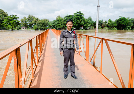 Poliziotto Thai permanente sulla luminosa arancione passerella metallica a allagata Ping diga sul fiume a sud di Chiang Mai nel nord della Thailandia Foto Stock
