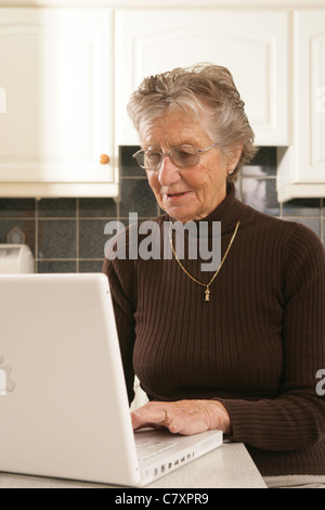 Una donna nella sua metà anni ottanta usando un computer portatile nella sua cucina. Foto Stock