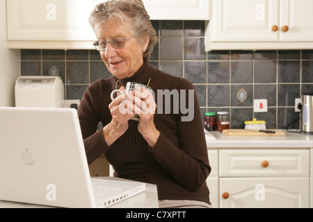 Una donna nella sua metà anni ottanta usando un computer portatile nella sua cucina. Foto Stock