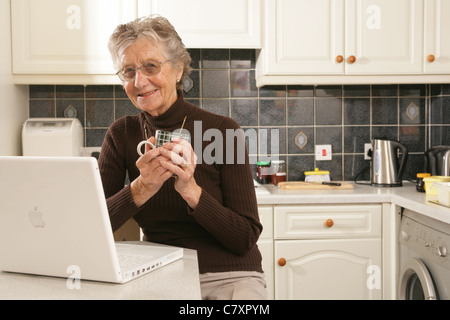 Una donna nella sua metà anni ottanta usando un computer portatile nella sua cucina. Foto Stock