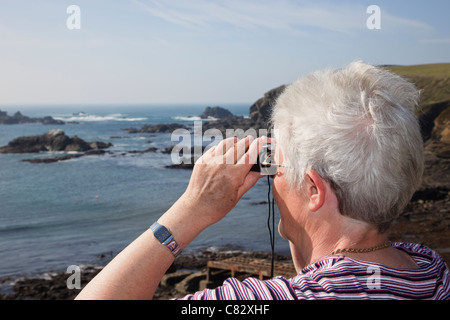 Senior donna pensionato guardando attraverso il binocolo al mare sulla costa di più a sud il punto sul territorio continentale del Regno Unito. Lizard Point Cornwall Inghilterra REGNO UNITO Foto Stock