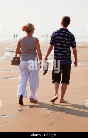 Coppia giovane camminando sulla spiaggia sabbiosa, l'uomo a piedi nudi con pantaloni arrotolato che trasportano le scarpe con la bassa marea. Cornwall Inghilterra England Regno Unito. Foto Stock