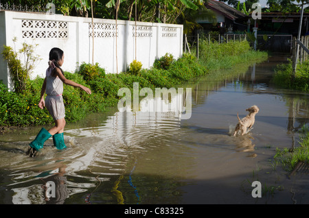 Giovane ragazza di indossare stivali di gomma passeggiate attraverso strada allagata con un piccolo cane che corre attraverso l'acqua di fronte a lei in Thailandia Foto Stock