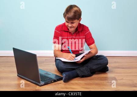 Foto di uno studente che sta facendo il suo dovere sabato sul pavimento guardando al suo portatile mentre la scrittura in un notebook Foto Stock