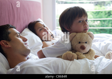 Ragazzo seduto nel letto con i suoi genitori e suo orsacchiotto Foto Stock