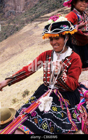 Pisac, Perù. Donna sorridente in abito tradizionale la filatura della lana e la tessitura di un cinturino utilizzando un telaio a pedale. Foto Stock