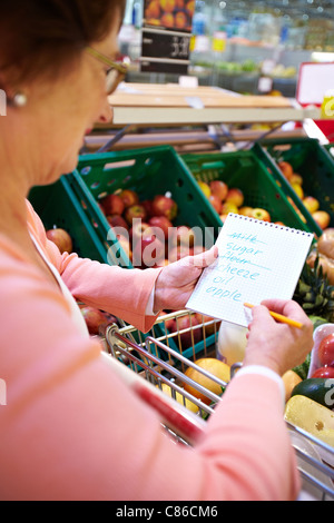 Immagine di senior donna che guarda la lista del prodotto con la merce nel carrello nelle vicinanze Foto Stock