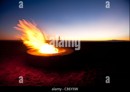 Un paio di passeggiate lungo una spiaggia dietro a un incendio di grandi dimensioni durante il tramonto. Foto Stock