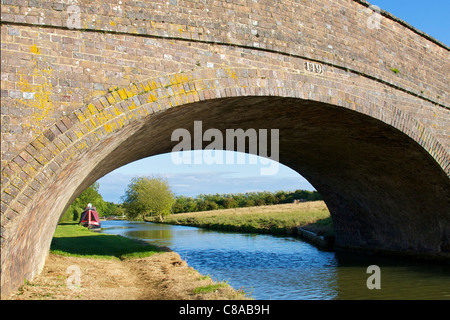 Narrowboat su un canale incorniciate da mattoni ponte arcuato e set contro il profondo blu del cielo dello sfondo. Un blocco può essere visto in distanza Foto Stock