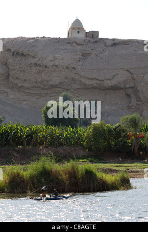 La sezione di lussureggiante verde fiume Nilo bank contro una roccia con il santuario in cima con piccole barche da pesca sul fiume di fronte, Egitto Foto Stock