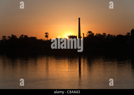 Una sezione del fiume Nilo banca con il sole che tramonta in un bagliore dorato dietro camini industriali riflessi nell'acqua, Egitto, Africa Foto Stock