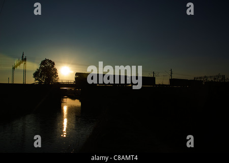 Una classe 66 locomotive diesel in silhouette attraversa il canale di Coventry e il fiume Anker a Bushmills con un trasporto intermodale Foto Stock