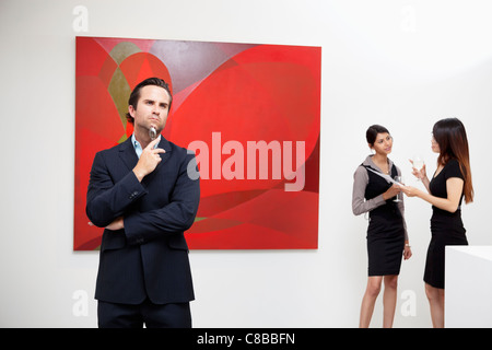 Giovane uomo di pensare con due donne parlando in background in galleria d'arte Foto Stock