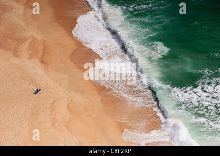 Nazaré beach, un solitario sunbather orologi di grandi onde infrangersi sulla riva, da sopra al Sitio Foto Stock