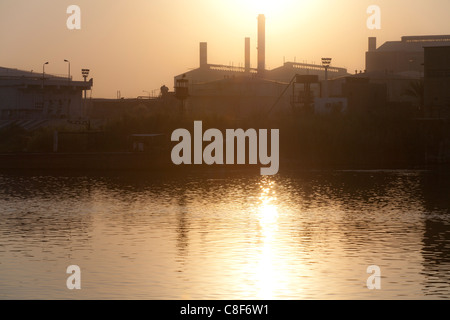 Una sezione del fiume Nilo banca con il sole che tramonta in un bagliore dorato dietro camini industriali riflessi nell'acqua, Egitto, Africa Foto Stock