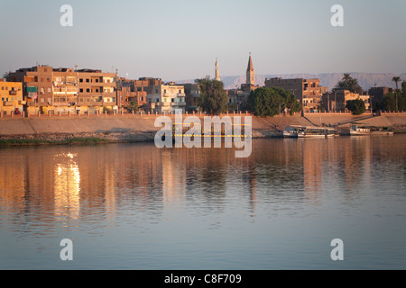 Una sezione del fiume Nilo banca con piccola città riflettendo in acqua ancora con golden dawn luce, con la Chiesa Copta e moschea Foto Stock