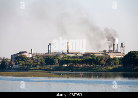 Una sezione del fiume Nilo banca con impianto industriale con comignoli fumanti dietro alberata in banca e numerose piccole barche da pesca Foto Stock