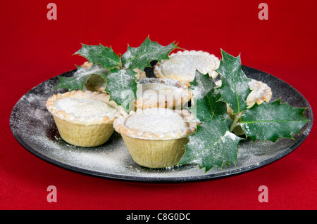 Piatto di carne macinata torte sulla piastra nera con holly ciuffi contro una festosa sfondo rosso Foto Stock