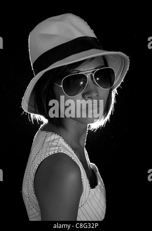 Una immagine illuminata da dietro di una bella ragazza asiatica indossando occhiali da sole e cappello. L'immagine è in bianco e nero. Foto Stock