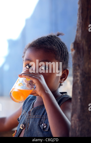 Una giovane ragazza malgascia con un taglio di capelli tradizionale di bere soda arancione al di fuori in una spianata in Hell-Ville, Nosy Be, Madagascar Foto Stock