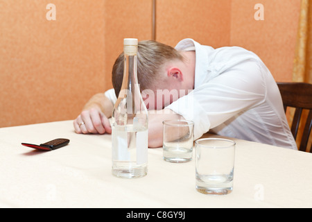 Ubriaco posa sul tavolo con una bottiglia di vodka e bicchieri di alcol. Sleeping ragazzo in maglia bianca Foto Stock