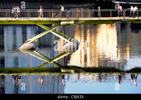 La gente camminare attraverso il nord passerella di Dock e la riflessione sulla West India Quay a Canary Wharf, Isle of Dogs Tower Hamlets, East London Inghilterra England Regno Unito Foto Stock