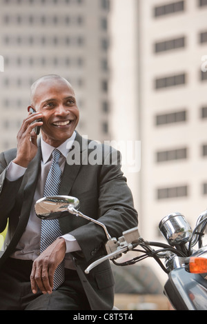 Imprenditore nero seduto su uno scooter parlando al cellulare Foto Stock