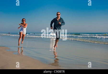 Coppia giovane 20s in esecuzione sulla sabbia bagnata sulla spiaggia cielo blu oceano occhiali da sole ridere per godersi la vita abbigliamento casual Foto Stock