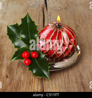 Masterizzazione decorativo rosso e oro candela con un rametto di agrifoglio fresco su uno sfondo di vecchio legno stagionato Foto Stock