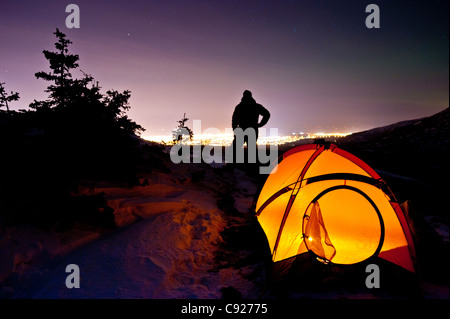 Silhouette di un uomo in piedi da una tenda a lume di candela in Chugach State Park con la città di ancoraggio in background, Alaska Foto Stock