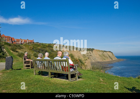 I turisti anziani che si trovano seduti sulla panca che guarda fuori Mare in estate Robin Hoods Bay North Yorkshire Inghilterra Regno Unito Gran Bretagna Foto Stock