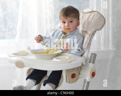 Un anno e mezzo di lavoro vecchio ragazzo seduto in una sedia alta e mangiare la minestra con un cucchiaio, riversandosi sulla sua maglietta Foto Stock