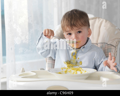 Simpatico bimbo seduto in una sedia alta e mangiare la minestra con un cucchiaio, riversandosi sulla sua maglietta Foto Stock