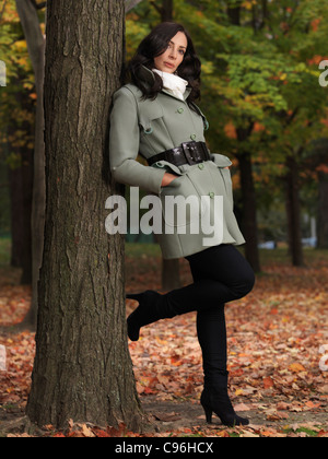 Vestiti alla moda per la stagione autunnale donna che indossa un manto verde, appoggiata contro un albero in un parco Foto Stock