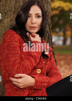 Bella donna cuddling fino in un maglione rosso. Appoggiata contro un albero in autunno. Foto Stock