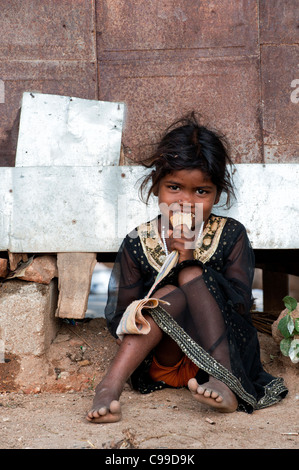 Povero indiano mendicante nomadi ragazza seduta contro una capanna di mangiare un biscotto. Andhra Pradesh, India Foto Stock