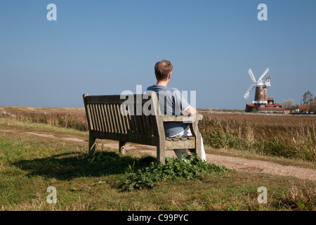 Uomo seduto su un banco di lavoro sul bordo delle paludi con Cley Windmill in background, Cley accanto al mare, Norfolk Foto Stock