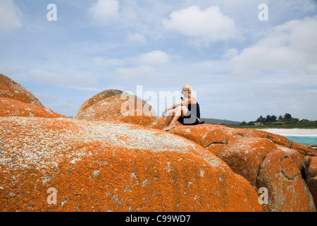 Una donna si siede in cima al lichene arancione rocce di Binnalong Bay nella baia di incendi. St Helens, Tasmania, Australia Foto Stock