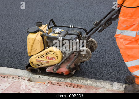Workman utilizzando una dynapac per compact asfalto (asfalto) lungo il bordo del marciapiede (cordolo) dopo il nuovo manto stradale era stata prevista. Foto Stock