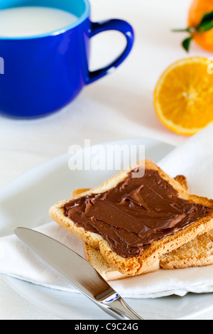 La prima colazione con cioccolato da spalmare sul pane tostato, latte e arancione Foto Stock