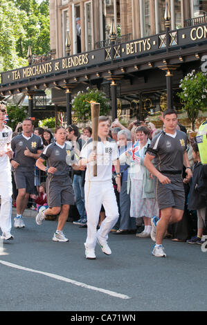 Giovane ragazzo adolescente agendo come Olympic Torchbearer (maschio allievo dalla scuola locale) è portatore di & holding torce fiammeggianti aloft, guardato da grande folla di ben wishers & accompagnata dalla torcia di sicurezza i membri del team in grigio kit in esecuzione. Al di fuori di Betty's Tea Rooms, Harrogate Town Center, nello Yorkshire, Inghilterra, Regno Unito. Martedì 19 Giugno 2012. Foto Stock