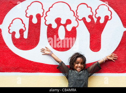 Sorridendo felice povero indiano mendicante nomadi ragazza in piedi di fronte a una gioia murale su una parete. Andhra Pradesh, India Foto Stock