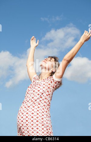 Donna incinta sorridente permanente all'aperto contro il cielo sereno le armi in aria e a basso angolo di visione Foto Stock