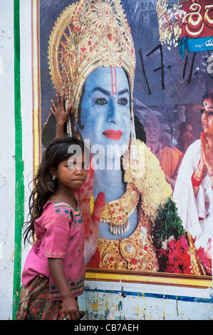 Povero indiano mendicante nomadi ragazza in piedi di fronte ad una Rama poster su un indiano street. Andhra Pradesh, India Foto Stock