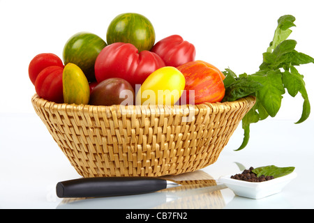 Due cesti con una varietà di pomodori organico con la lama di un coltello e grani di pepe in un piatto di fronte a uno sfondo bianco Foto Stock